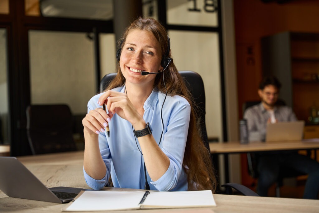 ALDB Kontakt: Lächelnde Mitarbeiterin mit Headset am Schreibtisch nimmt Anfragen per Telefon entgegen